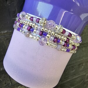 May include: Stack of beaded bracelets in shades of purple, silver, and clear. The bracelets feature a variety of beads, including small silver beads, larger purple beads, and crystal accents. The bracelets are stacked on a light purple, glittery surface.