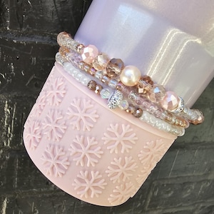 May include: A set of four beaded bracelets with pink, white, and brown beads. The bracelets are wrapped around a pink tumbler with a snowflake pattern.