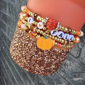 May include: A stack of beaded bracelets featuring metallic gold, orange, and peach beads, interspersed with white letter beads spelling I HEART FALL, a glittery orange bead, and a dangling orange pumpkin charm with a green stem.