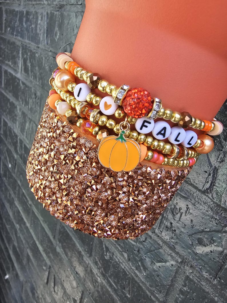 May include: A stack of beaded bracelets featuring metallic gold, orange, and peach beads, interspersed with white letter beads spelling I HEART FALL, a glittery orange bead, and a dangling orange pumpkin charm with a green stem.