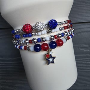 May include: A stack of beaded bracelets in red, white, and blue. The bracelets feature various bead sizes and textures, including sparkly red and silver beads. One bracelet has a star charm. The bracelets are displayed on a white cylindrical form.