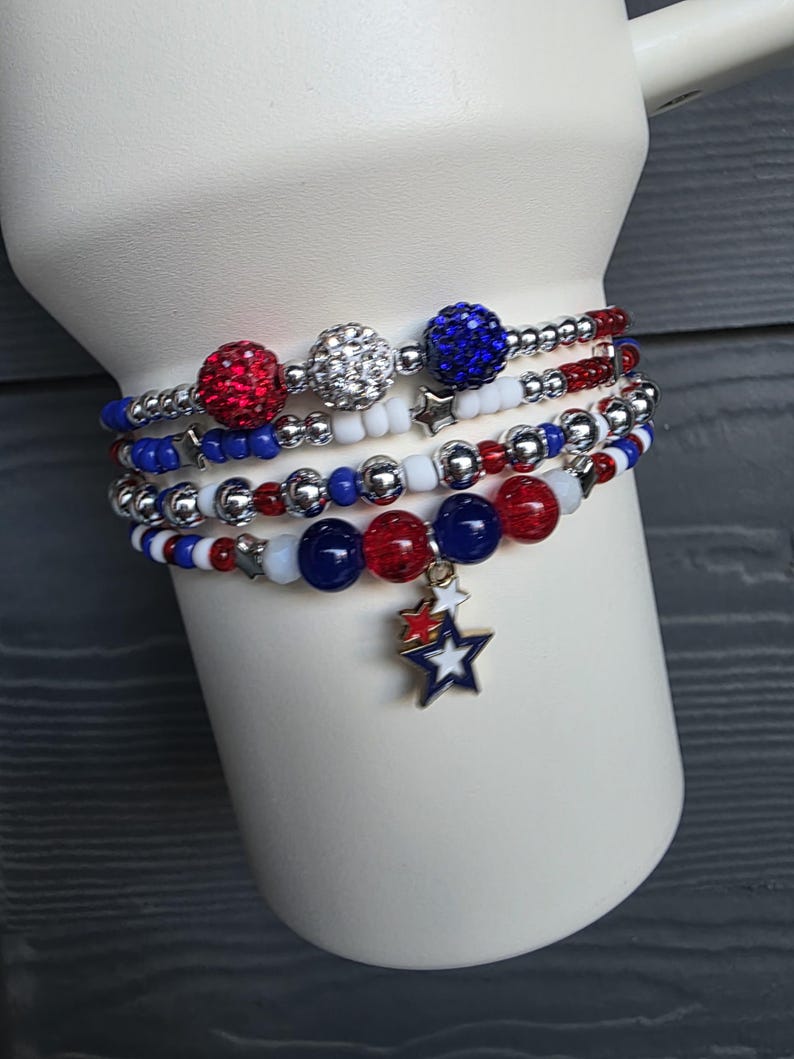 May include: A stack of beaded bracelets in red, white, and blue. The bracelets feature various bead sizes and textures, including sparkly red and silver beads. One bracelet has a star charm. The bracelets are displayed on a white cylindrical form.