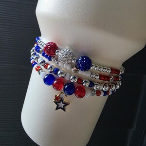 May include: A stack of beaded bracelets in red, white, and blue. The bracelets feature various bead sizes and textures, including sparkly and solid beads. A gold star charm hangs from one bracelet, adding a decorative touch.