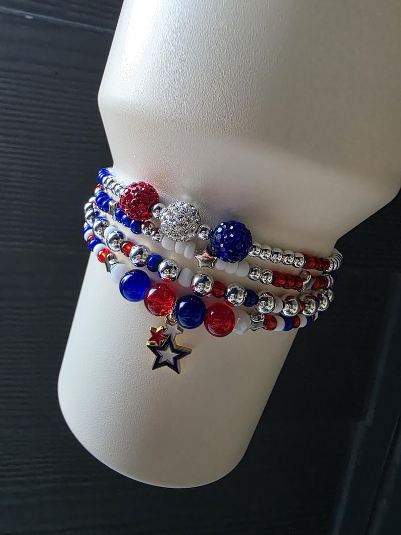 May include: A stack of beaded bracelets in red, white, and blue. The bracelets feature various bead sizes and textures, including sparkly and solid beads. A gold star charm hangs from one bracelet, adding a decorative touch.