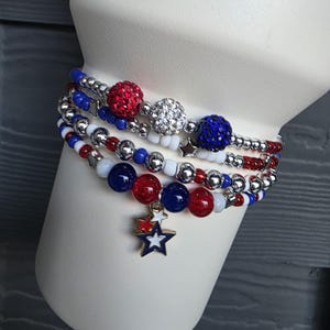 May include: A collection of beaded bracelets in red, white, and blue, displayed on a white form. The bracelets feature various bead sizes and textures, including sparkling red and silver beads. A star charm hangs from one bracelet.