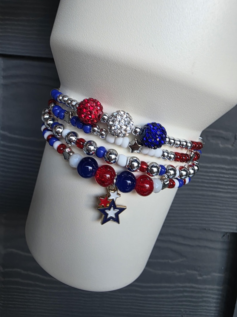 May include: A collection of beaded bracelets in red, white, and blue, displayed on a white form. The bracelets feature various bead sizes and textures, including sparkling red and silver beads. A star charm hangs from one bracelet.