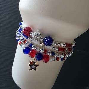 May include: A stack of beaded bracelets in red, white, and blue. The bracelets feature various bead sizes, including large, sparkling orbs and smaller beads. One bracelet has a gold star charm. The bracelets are displayed on a white form.