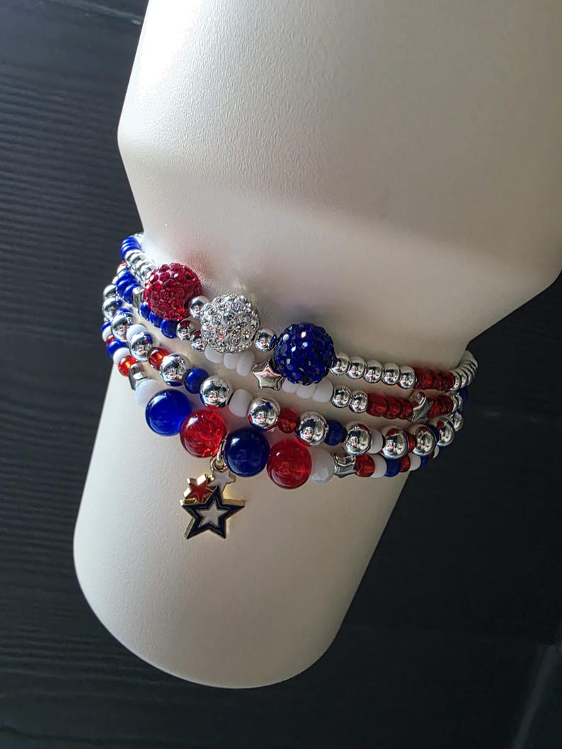 May include: A stack of beaded bracelets in red, white, and blue. The bracelets feature various bead sizes, including large, sparkling orbs and smaller beads. One bracelet has a gold star charm. The bracelets are displayed on a white form.