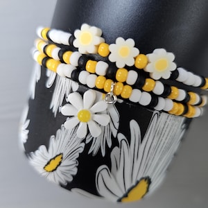 May include: A set of beaded bracelets with yellow and white beads and daisy charms. The bracelets are stacked on a black background with a daisy print.