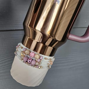 Könnte beinhalten: Ein roségoldener Reisebecher mit rosa Griff und weißer, strukturierter Basis. Die Basis ist mit mehreren Strängen aus rosa, goldenen und transparenten Perlen verziert, die einen dekorativen Akzent bilden.
