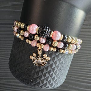 Könnte beinhalten: Drei Armbänder mit Perlen in Rosa, Schwarz und Gold. Ein Armband hat eine schwarze, kristallbesetzte Perle. Ein goldener Kronenanhänger hängt an einem Armband. Die Armbänder werden auf einer schwarzen, strukturierten Oberfläche präsentiert.
