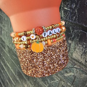 May include: A stack of beaded bracelets featuring gold, orange, and peach-colored beads. The bracelets include white letter beads spelling I HEART FALL, a central orange glitter bead, and a dangling orange pumpkin charm with a green stem.