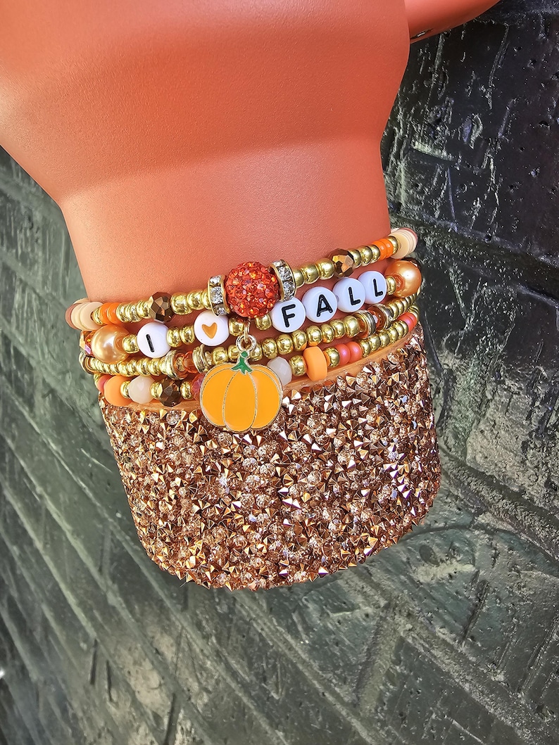 May include: A stack of beaded bracelets featuring gold, orange, and peach-colored beads. The bracelets include white letter beads spelling I HEART FALL, a central orange glitter bead, and a dangling orange pumpkin charm with a green stem.