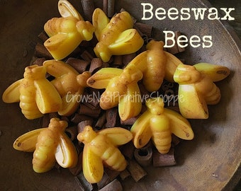 May include: A collection of yellow beeswax bee-shaped candles. The candles are arranged in a wooden bowl with cinnamon sticks. The text "Beeswax Bees" is visible in the upper right corner.