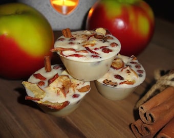 May include: Three small, cream-colored, round candles with dried apple and cinnamon decorations. Two red and green apples and a lit candle in a heart-shaped holder are in the background. Cinnamon sticks and peppercorns are on the wooden surface.