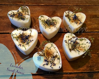 May include: Six heart-shaped white candles with dried lavender and other botanicals embedded in the wax. The candles are arranged on a wooden surface. The text "Lavender Sweet Annie" is visible in the lower left corner of the image.