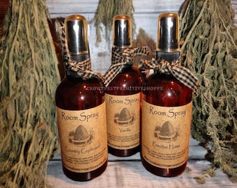 May include: Three brown glass bottles with black and white checkered ribbon tied around the neck. Each bottle has a brown paper label with the text "Room Spray" and a different scent: "Country Cupboard", "Vanilla", and "Primitive Home".