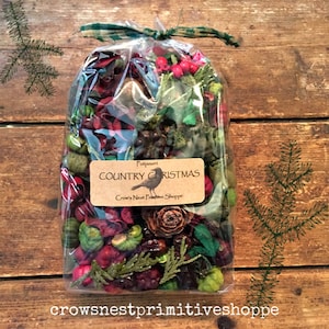 May include: A clear cellophane bag filled with potpourri. The potpourri includes red berries, green accents, and pine cones. A label reads "Country Christmas" with a bird illustration. The bag is tied with a green and white checkered ribbon.