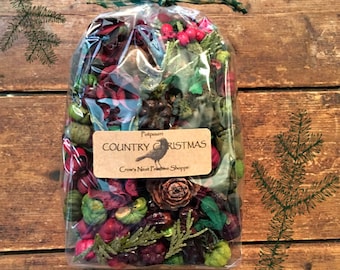 May include: A clear cellophane bag filled with potpourri. The potpourri includes red berries, green accents, and pine cones. A label reads "Country Christmas" with a bird illustration. The bag is tied with a green and white checkered ribbon.