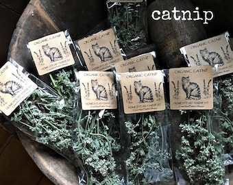 May include: Multiple clear plastic bags filled with dried green catnip. Each bag features a tan paper label with a black illustration of a cat, sprigs of lavender, and the text Organic Catnip, Homestead Harvest, Crows Nest Primitive Shoppe.