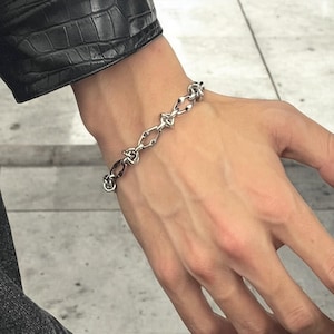 May include: A silver chain bracelet with a unique link design. The bracelet is worn on a person's wrist.