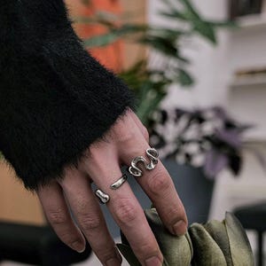 Può includere: Due anelli d'argento sulla mano di una persona. L'anello sull'indice è una fascia spessa e ondulata. L'anello sul medio è una fascia sottile e liscia.