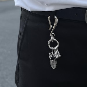 以下が含まれることがあります： 黒いベルトループに取り付けられたシルバーカラーのキーチェーン。キーチェーンは、装飾的なフック、ねじれたロープのデザインのリング、黒い石が付いた羽のペンダントが特徴です。鍵が見えます。
