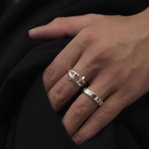Puede incluir: Dos anillos de plata se muestran en una mano. Uno presenta un diseño de barco con un elemento puntiagudo, mientras que el otro tiene una banda con pequeños detalles en relieve. Los anillos están sobre un fondo oscuro.