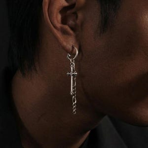 Op de afbeelding: Zilveren oorbel met een kruisvormige hanger en kettingdetail. De oorbel is bevestigd aan een ring die in het oor wordt gestoken. De kruishanger heeft ingewikkelde details.