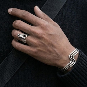 May include: A silver ring and bracelet set. The ring features a layered chevron design. The bracelet has a wavy, layered design. Both pieces are worn on a hand against a dark background. The jewelry is likely made of sterling silver.