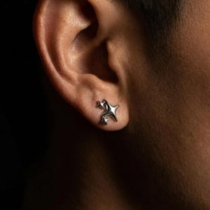 May include: A pair of silver star-shaped stud earrings. The earrings are worn on an ear, with a dark background. The earrings have a three-dimensional design, with multiple points and a shiny finish. The earrings are small and delicate.