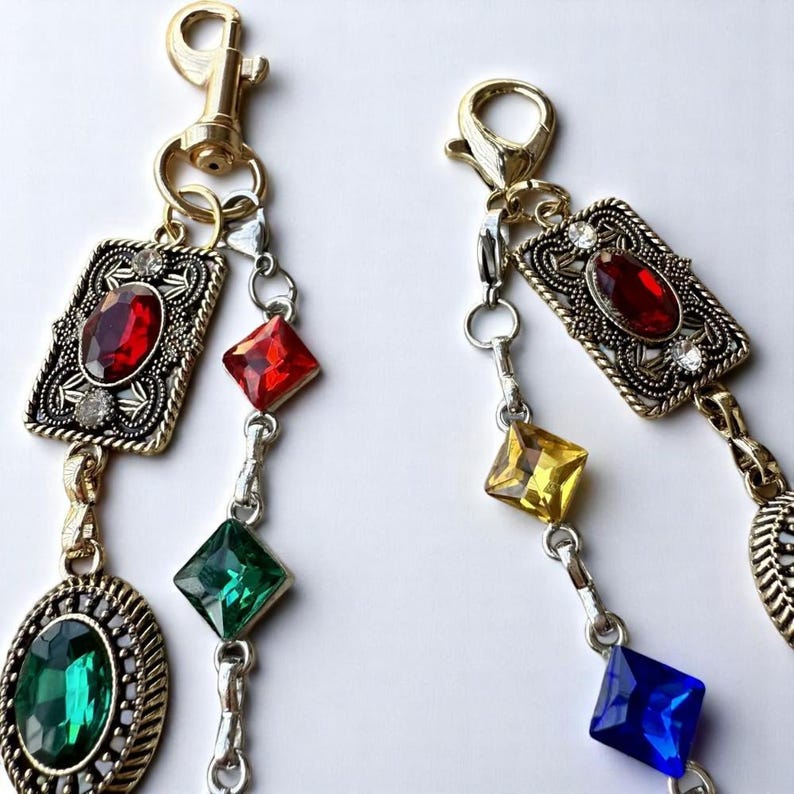 May include: Two gold keychains with a chain of colorful rhinestones. The keychains feature a gold clasp and a decorative, ornate frame with a red rhinestone in the center.