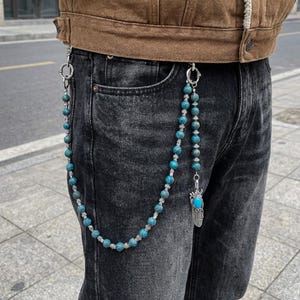 May include: A beaded chain with turquoise and silver accents hangs from a belt loop. The chain features round, blue-green beads and silver-colored spacers. A silver feather charm with a turquoise stone dangles from the chain.