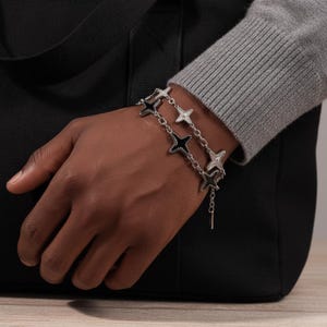 May include: A silver bracelet with star-shaped charms. The bracelet has two strands of linked stars, some with black enamel and others with clear crystals. The bracelet is worn on a wrist, with a black bag in the background.