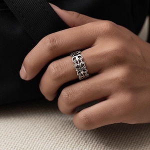 Puede incluir: Un anillo plateado con un diseño calado ornamentado con formas repetidas en forma de cruz. El anillo se lleva en un dedo, mostrando sus intrincados detalles y su acabado pulido. El diseño del anillo incorpora pequeños acentos brillantes.