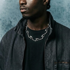May include: A silver-colored necklace with an abstract design is worn over a black t-shirt. The necklace is composed of interconnected, curved shapes. The person is wearing a black denim jacket with a zipper.