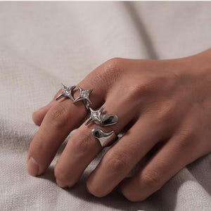 Könnte beinhalten: Nahaufnahme einer Hand mit vier silbernen Ringen. Zwei Ringe haben sternförmige Designs mit klaren Edelsteinen, während die anderen beiden abstrakte, fließende Formen aufweisen.