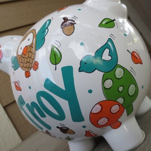 Może przedstawiać: Biała ceramiczna skarbonka z malowanym wzorem w kolorze turkusowym, zielonym, pomarańczowym i brązowym. Skarbonka ma namalowane imię "Troy".