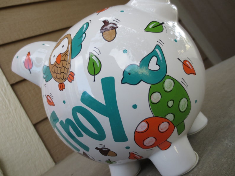 Może przedstawiać: Biała ceramiczna skarbonka z malowanym wzorem w kolorze turkusowym, zielonym, pomarańczowym i brązowym. Skarbonka ma namalowane imię "Troy".