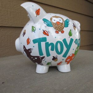 Może przedstawiać: Biała ceramiczna skarbonka z motywem leśnym. Skarbonka jest ozdobiona brązową sową, jeleniem, liśćmi, żołędziami i grzybami. Imię "Troy" jest napisane na boku turkusowymi literami.
