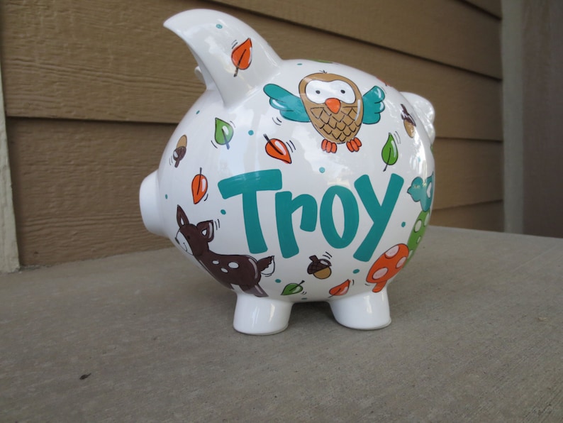 Może przedstawiać: Biała ceramiczna skarbonka z motywem leśnym. Skarbonka jest ozdobiona brązową sową, jeleniem, liśćmi, żołędziami i grzybami. Imię "Troy" jest napisane na boku turkusowymi literami.