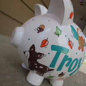 Może przedstawiać: Biała ceramiczna skarbonka z wzorem leśnym w kolorze turkusowym, brązowym i pomarańczowym. Skarbonka ma namalowanego jelenia, sowę i liście. Imię "Troy" jest napisane turkusowym kolorem z boku.