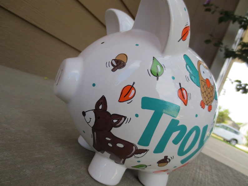 Może przedstawiać: Biała ceramiczna skarbonka z wzorem leśnym w kolorze turkusowym, brązowym i pomarańczowym. Skarbonka ma namalowanego jelenia, sowę i liście. Imię "Troy" jest napisane turkusowym kolorem z boku.