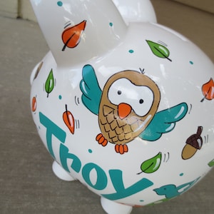 Może przedstawiać: Biała ceramiczna skarbonka z ręcznie malowanym wzorem przedstawiającym sowę, liście i imię "Troy" w kolorze turkusowym.