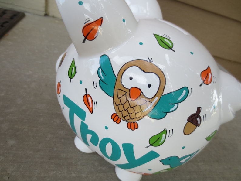 Może przedstawiać: Biała ceramiczna skarbonka z ręcznie malowanym wzorem przedstawiającym sowę, liście i imię "Troy" w kolorze turkusowym.