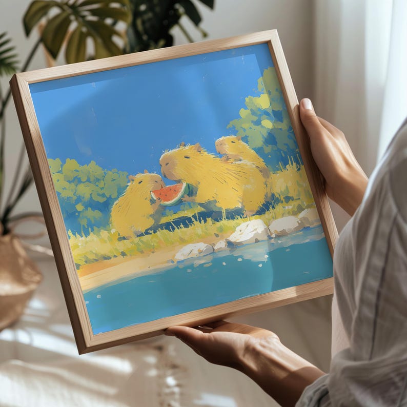 May include: A square wooden frame containing a colorful illustration of three yellow capybaras near a blue body of water. One capybara is eating a slice of red watermelon. The scene includes green foliage, white rocks, and a bright blue sky.