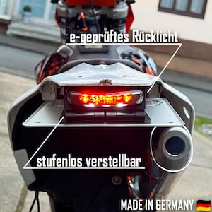 May include: A red motorcycle taillight with the text "e-geprüftes Rücklicht" and "stufenlos verstellbar" on the image. The taillight is mounted on a motorcycle with a black exhaust pipe. The text "MADE IN GERMANY" is also visible on the image.
