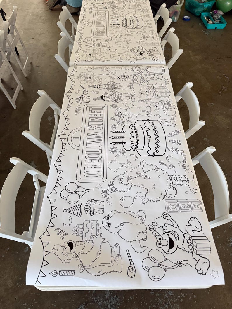 May include: A long white paper coloring table runner featuring black line art illustrations of Sesame Street characters, birthday cakes, balloons, and gift boxes. The text OREOLUWA STREET is printed on a sign graphic on the paper surface.