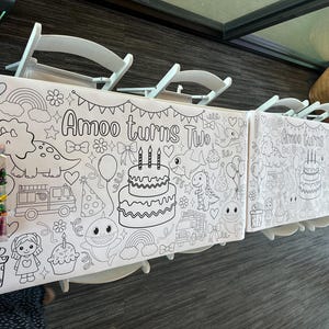 May include: A long white paper coloring tablecloth featuring black line art illustrations of a two-tier birthday cake, a dinosaur, a fire truck, a shark, rainbows, and the text "Amoo turns Two". A clear plastic container filled with colorful crayons sits on top.