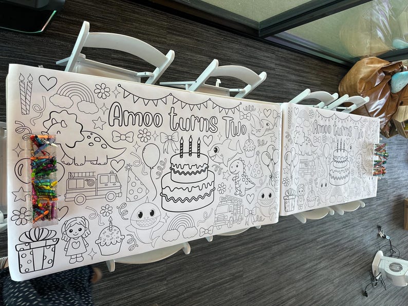May include: A long white paper coloring tablecloth featuring black line art illustrations of a two-tier birthday cake, a dinosaur, a fire truck, a shark, rainbows, and the text "Amoo turns Two". A clear plastic container filled with colorful crayons sits on top.